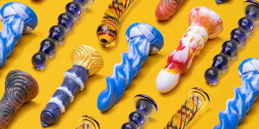 Varios dildos de vidrio y cerámica en colores vibrantes sobre fondo amarillo, con diseños artísticos y texturas para diferentes tipos de estimulación.