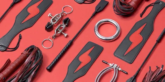 Conjunto de juguetes BDSM sobre fondo rojo, incluyendo látigos, fustas, paletas y pinzas para juegos de dominación.